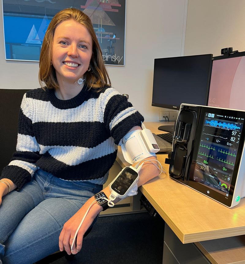 Marjolein Haveman zit aan tafel, met een draagbare computer aan haar arm, die verbonden is met haar vinger en een scherm waarop haar hartslag te zien is. 